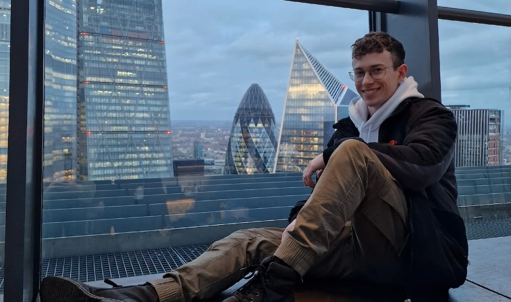 Simon Mayr sitzt entspannt in einem Hochhaus in London. Im Hintergrund sind bekannte Gebäude wie die „Gherkin“ und der „Cheesegrater“ zu sehen.